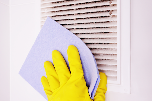 Homeowner wearing yellow cleaning gloves clean dirty air vents with cloth.