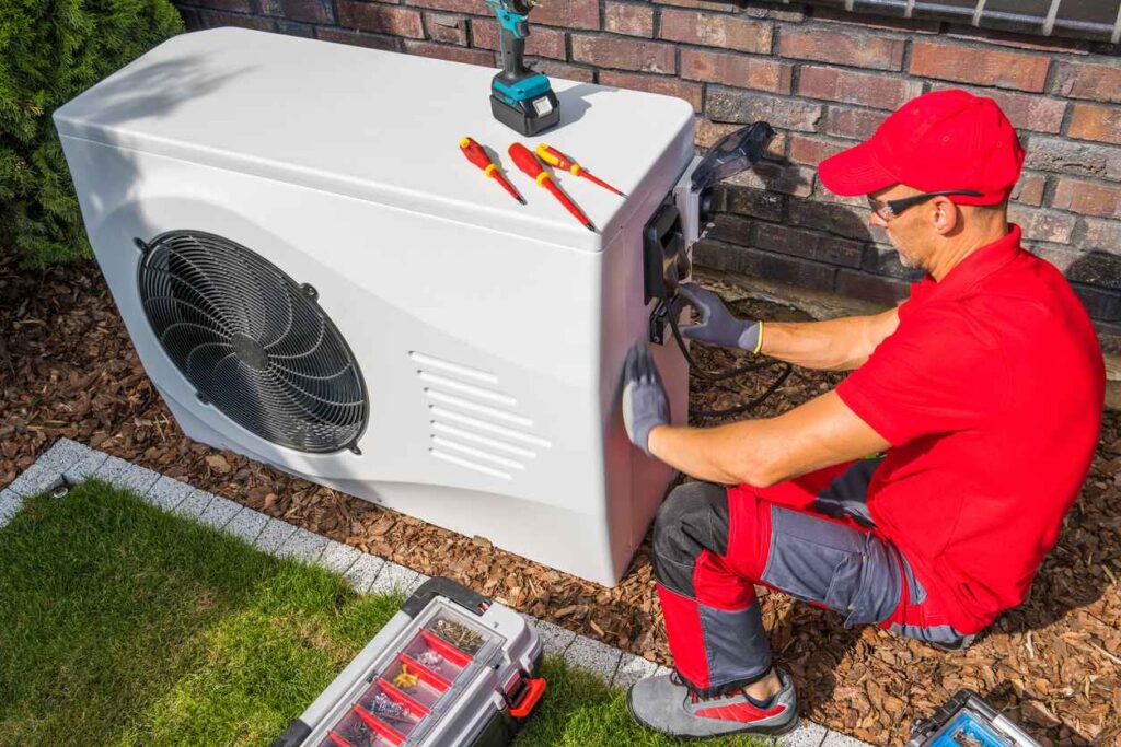 HVAC technician in a red shirt and red hat servicing a heat pump outside of a residential home